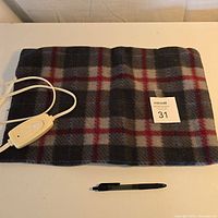 Full view of the small electric heating pillow on a white surface with a black pen for scale, showing the plaid fabric cover and attached power cord.