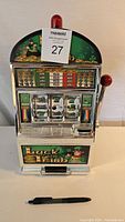 Front view of Luck of the Irish themed small slot machine showing lever, reels, coin slot, and green graphics with red light on top. Pen in front for size scale.