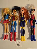 Photo of four Barbie dolls dressed as superheroes: Supergirl, Wonder Woman, Batgirl, and Harley Quinn, in used condition.