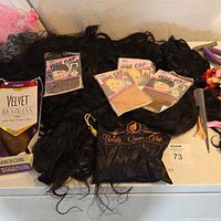 Lot of multiple wigs including Outre Velvet Brazilian Romanch Curl boxed wig, several loose black wigs, multiple wig caps in packaging, Vanity Queen Hair bag and hair tools gathered on white surface.