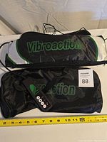 Photograph showing the Vibroaction massager in black and gray color with green logo, placed on a white surface with a tape measure indicating about 16 inches in length. Includes a black carrying bag with Vibroaction logo.