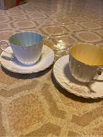 Photo showing two bone china tea cups and saucers, one with yellow interior, the other with green interior, placed on a patterned surface.