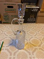 Bohemian crystal decanter with stopper visible in place, showing handle and cut glass design on a tiled floor with background boxes.