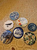 Seven collectors plates arranged on a table showing various designs including birds, flowers, horses, and nature scenes.