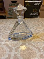 Crystal decanter sitting upright on a tiled floor, showing its triangular, faceted shape and reflective clear crystal material.