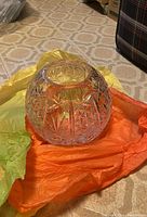 Photo of clear round cut crystal pot on orange and yellow tissue paper on patterned floor.