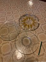 Three glass plates/platters on floor showing different patterns: bubbled texture plate, woven pattern plate, and gold starburst design plate.