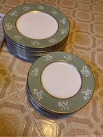 Two stacks of plates on the floor against a beige tiled background, showing the green rims, gold trim, and white floral decorations. Larger plates form one stack and smaller plates another.