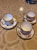 Photo showing three white china tea cup and saucer sets with gold rims and floral decorations, two Salisbury and one Jason set.