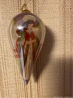 Front view of the teardrop-shaped glass ornament depicting an angel in robes with a staff, hanging on a textured fabric background.