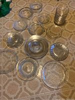 Top-down view of various clear crystal plates and bowls arranged on a patterned tablecloth. Plates and bowls vary in size and style, including textured and patterned designs.