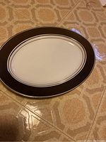 Oval platter with white center and dark border featuring thin double line detail near the inner edge, photographed on a tile floor.