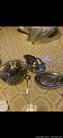 Photo of the silver tea set displaying ornate bowl, gravy boat, slotted spoon, and three platters on a patterned tablecloth.