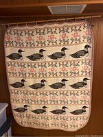 Front view showing the full size of the decorative vintage wall carpet with repeated patterns of stylized black ducks and pink flowers on cream background hanging from a rod.