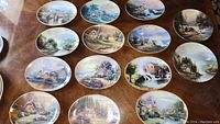 Photo showing 12 collectible Thomas Kinkade decorative plates arranged on a wooden surface. Plates are a mix of oval and round shapes with various landscape paintings.