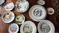 Overview of 14 collectible decorative plates arranged on a wooden table, showing various designs including nature, floral, and scenic themes