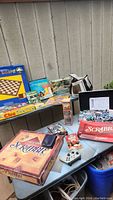 Photo showing a table with two Scrabble board game boxes, a Clue board game box, a poker chips box with chips, croquet set box, and various other game accessories including card cases and dice.