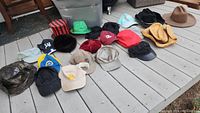 Wide shot of 15 assorted hats and caps laid out on a wooden floor, showing different styles and colors including black, red, beige, blue, brown, and a striped top hat.