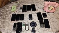 Photo of 17 phones including multiple smartphones, feature phones, and a wireless charger. Devices vary in brand and condition, many with cracked screens.