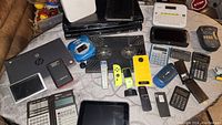 Overview photo showing various tech devices including calculators, label makers, remotes, and handheld electronic gadgets arranged on a table.