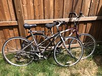 Two adult bicycles side by side leaning on a wooden fence outside on grass. One has silver frame with rear rack; other has burgundy front fork.