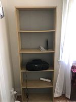 Photo of a 6ft light brown particle board bookshelf with five shelves in a corner near a window and an electrical outlet.