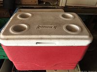 Photo showing front and top view of Coleman cooler with red body and white lid with 4 cup holders and Coleman logo.