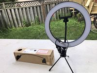 The web light set up on a white table outdoors with its original box beside it, showing the full setup including tripod legs and central phone holder.