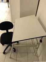 White drafting table with adjustable tilt and black office chair with wheels shown side by side on tiled floor