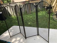 Three-panel folding metal fireplace screen standing outdoors on a white surface, showing the mesh and frame details.