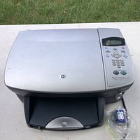 Full top and front view of HP PSC 2175 printer on white surface with green grass background. Printer shows scratches and includes one ink cartridge in plastic bag.