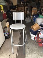 Front angle photo of IKEA white bar height stool with rectangular backrest and round seat, placed in a garage with assorted items around.