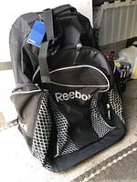 Front and side view of the large black Reebok wheeled hockey bag, highlighting the logo, zippered pockets and mesh panels.
