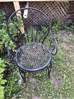 Front view of black metal patio chair showing rounded back with scrollwork and mesh seat, placed on grass outdoors.