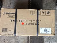 Photo of closed original cardboard box containing a StairMaster Twistlock Dumbbell. The box has the brand name, weight info and handling icons visible.