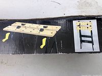Box featuring image of folding workbench with yellow adjustable pegs and black legs in folded and unfolded states.
