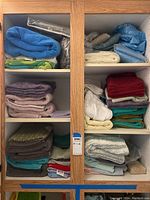 Wooden shelving cupboard containing stacks of assorted bath towels, bathmats, and folded curtains in multiple colors and fabrics.