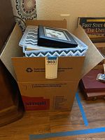 Cardboard box filled with books, some framed items, and covered with a textile.