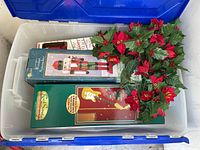 Blue plastic bin containing boxed musical nutcracker light, boxed guitar string lights, artificial holly garland with red flowers.