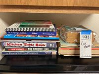 Stack of assorted cookbooks and a container with recipe cards on dark surface, wooden shelf behind