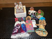 Collection of seven dolls displayed together on black surface, including cloth, woven and figurine types