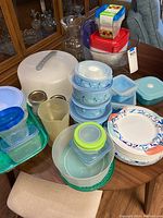 Photo showing a variety of plastic food storage containers, bowls, plastic cake carrier, disposable paper plates, and glass containers on a wooden table.
