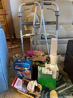 Photo of various home health aids including an aluminum walker, folding cane, boxed blood pressure monitor, hand weights, and other medical/personal care items on a carpeted floor.
