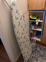 Ironing board with leaf patterned cover leaning against the wall near a wooden cupboard with household products inside.