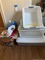 Multiple white plastic storage bins stacked including large bin with lid, medium holey bin, small bin with grey handles, and white wire tray. Miscellaneous office supplies visible to side.