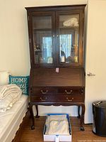 Front view of dark wood secretary desk with glass-front cabinet doors, brass knobs, drop front surface closed, and four drawers underneath.