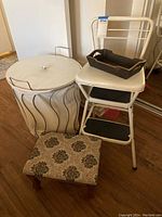 Photo showing white metal stool chair with padded seat, white hamper with wavy black lines, black tray on stool, and floral upholstered foot stool on floor.