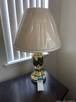Full view of brass colored table lamp with cream pleated lampshade on top, sitting on dark wooden table.