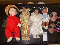 Full group photo showing four dolls and two New Orleans ceramic masks arranged on a dark surface, highlighting the variety in styles and sizes.