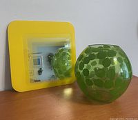 Yellow IKEA square mirror with rounded edges and reflective glass center next to green rounded glass lamp with leaf decoration on wooden surface.